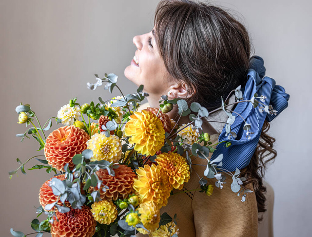 Young woman in a yellow dress with a blue ribbon in her hair, holding a bouquet of yellow and orange chrysanthemums, gray background. - Photo, Image