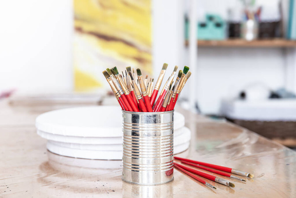 Red paint brushes in a metal can on a table with round canvases for painting against a blurred background. - Photo, Image