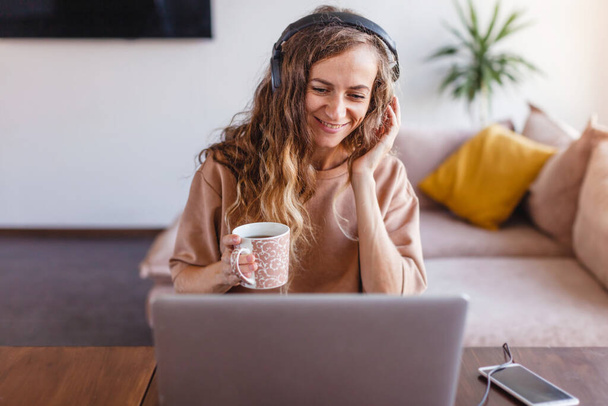 Woman is studying online video chat on a laptop. Happy female freelancer works on a remote house. Girl in headphones communicates by video call - Fotó, kép