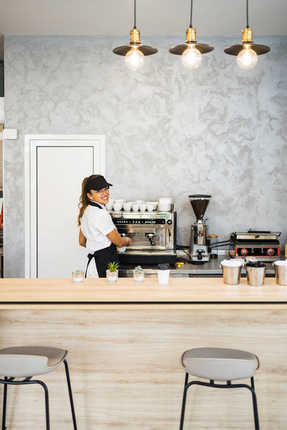 Beautiful and happy young female worker working in a bakery or fast food restaurant and using coffee machine. Positive people in small business concept. - Foto, Bild