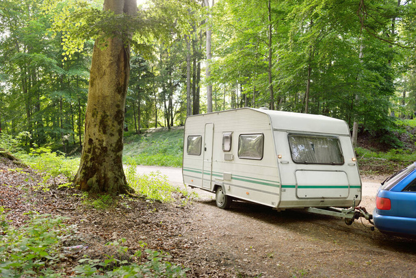 White caravan trailer and a car parked on the rural road under the mighty trees. Forest campsite. Europe. Lifestyle, freedom, travel, ecotourism, journey, recreation, transportation, RV, motorhome - Photo, Image