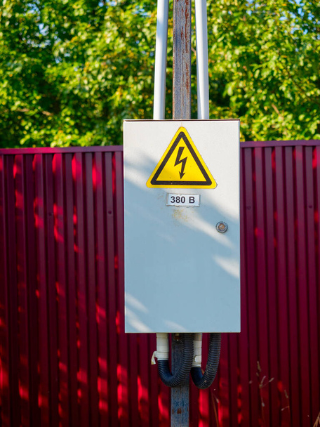 Közelkép egy szürke fém elektromos pajzsról, ami egy rúdon lóg. Figyelmeztető jel. Piros kerítés a háttérben - Fotó, kép