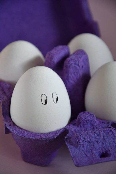 Photo of funny eggs with painted faces in a purple carton, preparation for Easter - Photo, Image