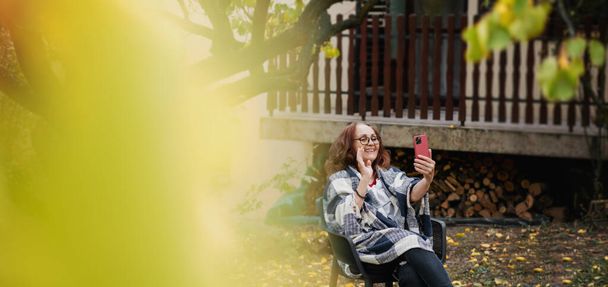 Beautiful mature happy smiling woman chatting using smartphone online in autumn garden of country house - Foto, Bild