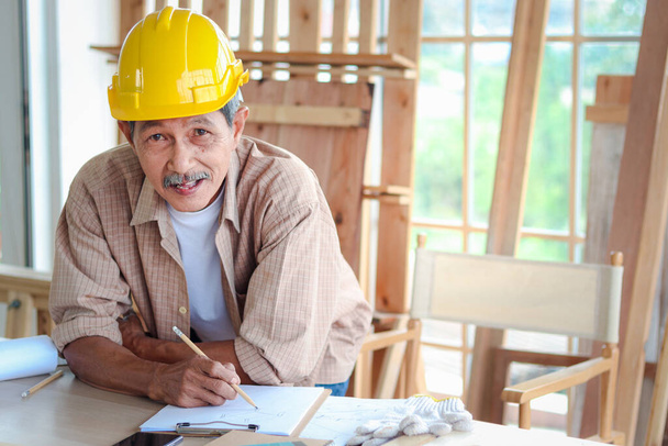 Portrait of smiling elderly carpenter working at wooden desk with different craft tools, enjoying his DIY hobby, senior craftsman drawing a design of piece of wood in carpentry woodwork workshop.  - Photo, Image