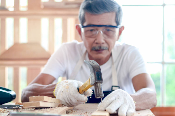 Starszy stolarz w okularach ochronnych gogle trzymający młotek i mocujący gwóźdź na desce, starszy rzemieślnik pracujący przy drewnianym biurku w stolarni ciesielskiej, cieszący się swoim hobby - Zdjęcie, obraz