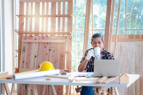 Carpinteiro asiático idoso com bigode fazendo uma pausa e bebendo um café, relaxando o artesão sênior na mesa de trabalho na oficina de carpintaria - Foto, Imagem