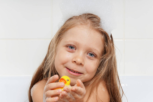 A cute, beautiful girl with long hair plays in the bathroom with a yellow rubber duck. Children's emotions in the bathtub with foam. Children's hygiene. Water games. Baby in the bathroom. The child washes the toy. - Foto, imagen