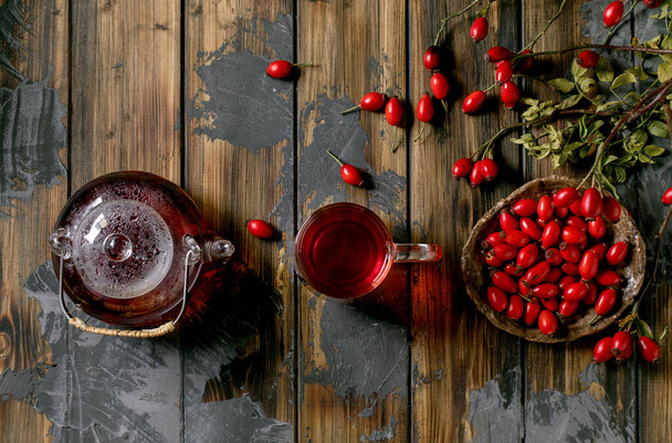 Rose hip berries herbal tea in glass teapot and cup standing on old wooden plank background with wild autumn berries around. Winter hot beverage. Flat lay, copy space - Fotografie, Obrázek