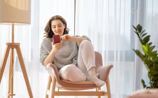 Happy casual beautiful woman is using a phone sitting on armchair at home. - Zdjęcie, obraz