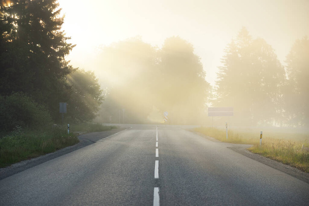 Une autoroute vide (route asphaltée) à travers les champs et la forêt dans un brouillard au lever du soleil. Paysage atmosphérique. Scène rurale estivale idyllique. Faucille météo, conduite dangereuse, road trip, concepts de vacances - Photo, image
