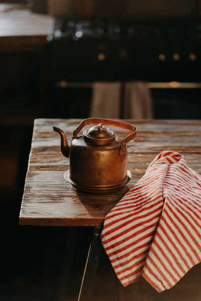 Retro aluminium kettle on wooden table with red striped towel near. Copper old teapot uses for making tea. Old fashioned kitchenware - Photo, Image