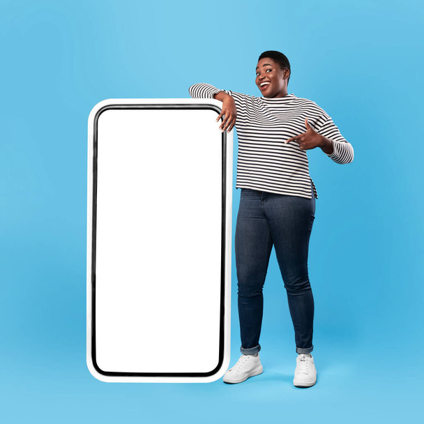 Negro hembra mostrando la pantalla de pie cerca de enorme Smartphone, fondo azul - Foto, imagen