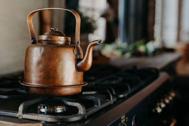 Bronze kettle in modern kitchen. Old vintage teapot on gas stove. Preparing tea. Aluminium teakettle. Sunny daylight from window. - Photo, image