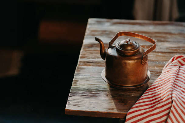 Старый медный металлический чайник на деревянном столе в темной комнате. Красное полосатое полотенце поблизости. Антикварный чайник для приготовления чая или кофе. Кулинарное оборудование - Фото, изображение