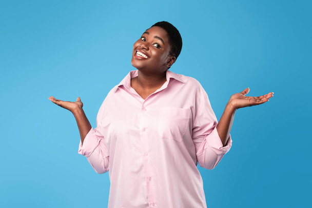 Positivo Oversized mulher negra encolhendo ombros posando sobre fundo azul - Foto, Imagem