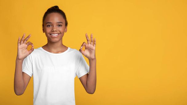 Χαρούμενη χαρούμενη έφηβη Αφρο-Αμερικάνα με λευκό t-shirt που δείχνει εντάξει σημάδι με τα χέρια - Φωτογραφία, εικόνα