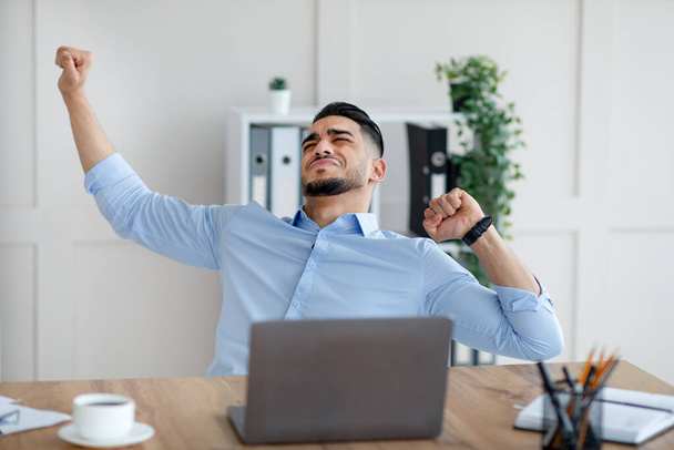 Millennial Άραβας επιχειρηματίας τεντώνει τους πονεμένους μυς του στο χώρο εργασίας, κάθεται στο γραφείο με φορητό υπολογιστή στο σύγχρονο γραφείο - Φωτογραφία, εικόνα