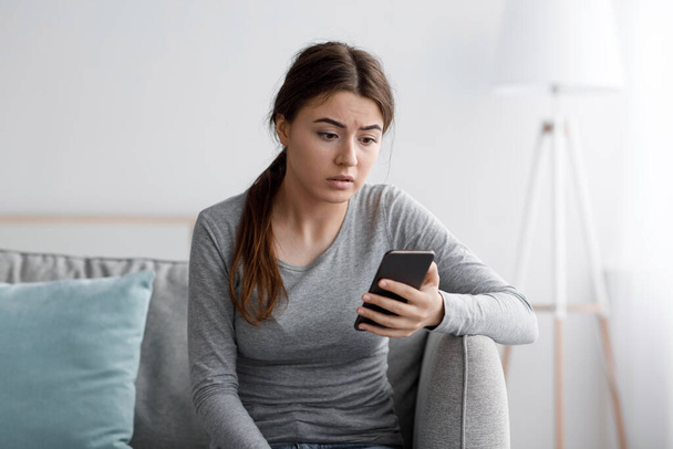 Sad young female sits on sofa look at smartphone and reading message at home, frustrated by received notification - Foto, Imagen