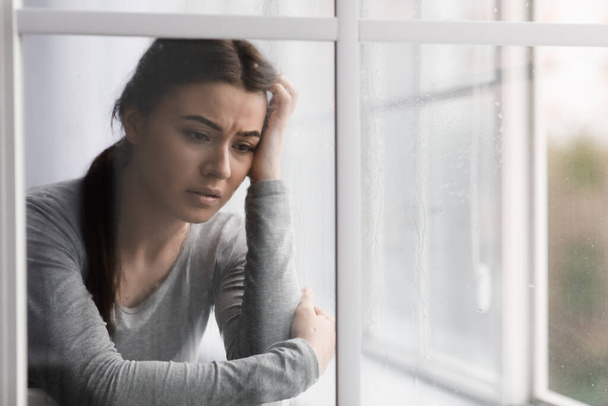 Δυστυχισμένη, λυπημένη Ευρωπαία κοπέλα που υποφέρει από κατάθλιψη, άγχος και διανοητικά προβλήματα κοντά στο παράθυρο με σταγόνες βροχής - Φωτογραφία, εικόνα