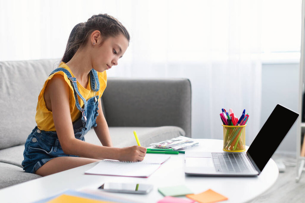 Egy lány ül az asztalnál, laptopot használ, tankönyvet ír. - Fotó, kép
