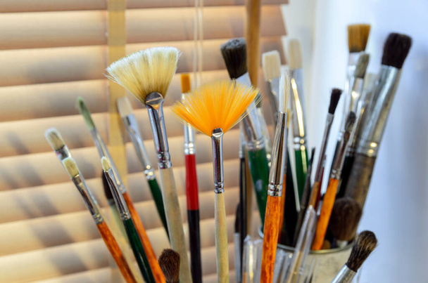 A set of used art brushes for painting and watercolors close up on the background of closed blinds  - 写真・画像