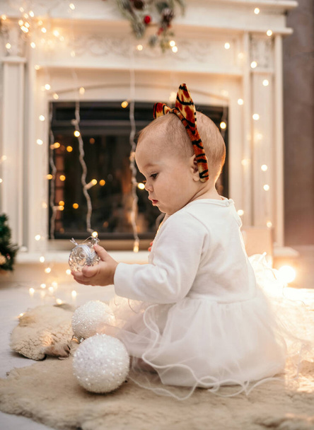 egy fehér kislány bolyhos fehér ruhában és egy fejpánt tigris fülekkel ül vele egy szobában a kandalló közelében karácsonyfa játékokkal - Fotó, kép
