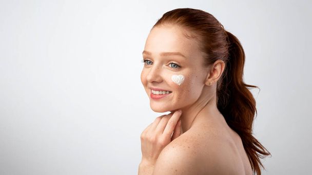 Happy Adolescent Girl Posing With Heart-Shaped Moisturizer On Face, Studio - 写真・画像