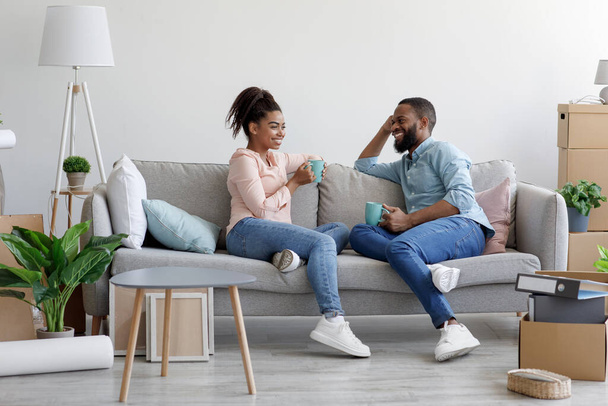 Boldog, csinos, ezeréves afro-amerikai család ül a kanapén és forró kávét iszik az új lakásban kartondobozokkal. - Fotó, kép