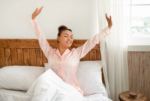Feliz y relajado después de una buena noche de sueño. Joven mujer afroamericana en pijama rosa estirándose después de despertar - Foto, Imagen