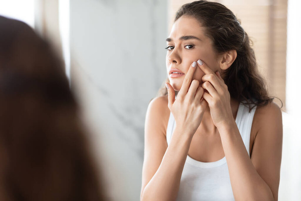 Unhappy Female Squeezing Pimples On Cheek Looking At Mirror Indoor - Photo, Image