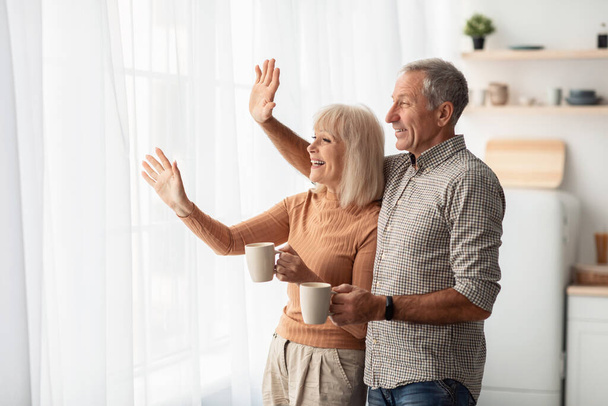 Happy Senior Couple Waving Hello Looking Out Of Window Indoor - Photo, Image