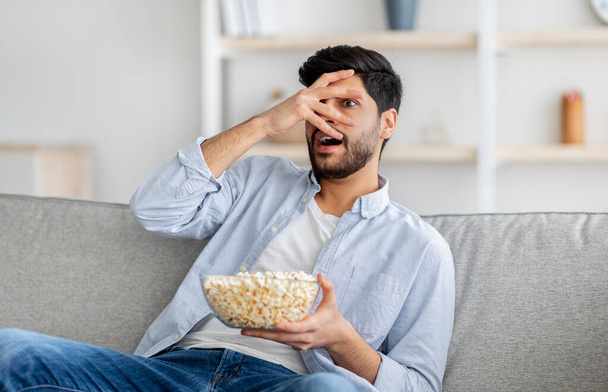 Young arab man watching scary movie, closing eyes in horror, eating popcorn, resting on sofa at home, free space - Photo, image
