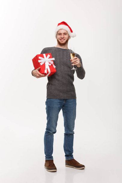 Weihnachtskonzept - glücklicher junger Mann mit Bart hält Geschenk in der Hand und Champagner feiert Weihnachten isoliert auf weißem Hintergrund. - Foto, Bild