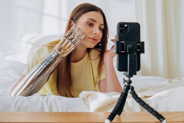 Bloggerin mit bionischer Armprothese führt eine Online-Sendung oder zeichnet ein Thema auf oder kommuniziert mit Freunden per Videoanruf, während sie auf einem Bett liegt - Foto, Bild