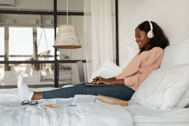 Side-view van Afro-Amerikaanse dame met behulp van laptop in de slaapkamer - Foto, afbeelding