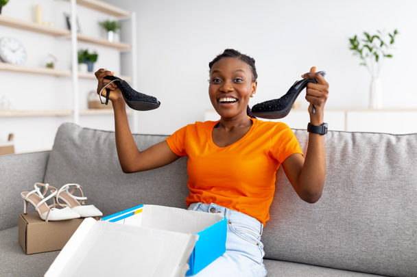 Zufriedenes Kundenkonzept. Positive schwarze Frau packt Karton aus, holt neue Schuhe aus Paket zu Hause - Foto, Bild