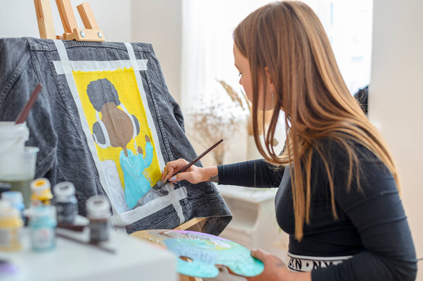 Artysta rysujący modę na jeansowej kurtce. Słoneczny warsztat krawiecki z farbami akrylowymi i sztalugami - Zdjęcie, obraz