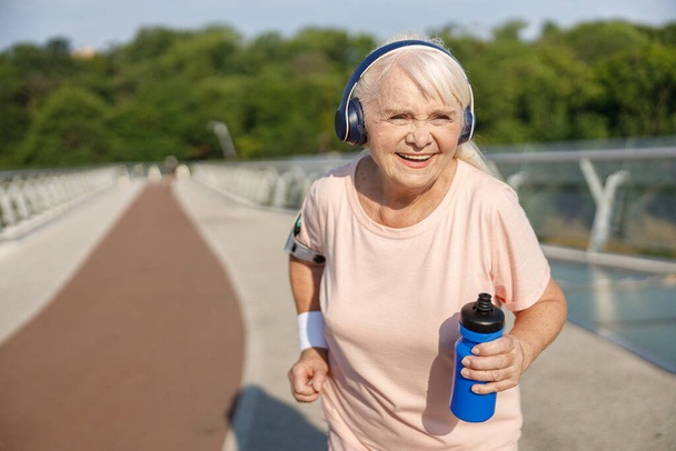 Señora mayor positiva con auriculares corre a lo largo del puente en verano - Foto, imagen