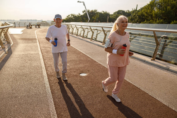 Cheerful blonde senior woman with husband run together along footbridge at sunset - Photo, Image