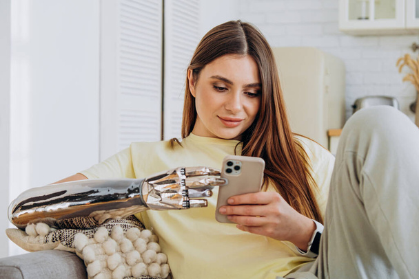 A happy woman with a bionic prosthetic arm sits on the couch with a phone and communicates online with friends or surfs the Internet or makes online purchases - Photo, Image
