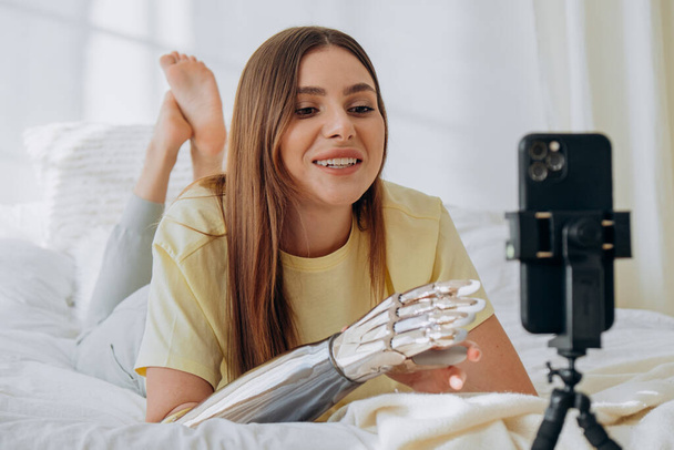 Γυναίκα blogger με ένα βιονικό προσθετικό χέρι οδηγεί μια διαδικτυακή εκπομπή ή καταγράφει ένα θέμα ή επικοινωνεί με φίλους με βιντεοκλήση ενώ βρίσκεται ξαπλωμένη στο κρεβάτι - Φωτογραφία, εικόνα