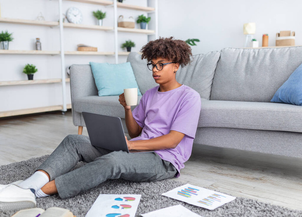 Чернокожий студент-подросток делает образовательный проект для онлайн-курса, пьет кофе дома - Фото, изображение