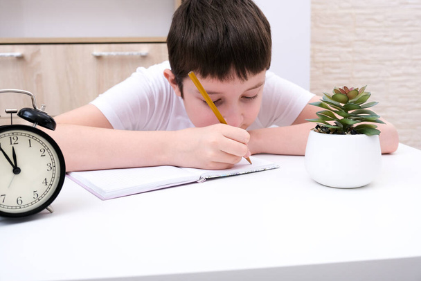 A concentrated boy writing in a textbook, exercisebook, a schoolboy doing his homework at home, an alarm clock in front of hin showing deadline. - Фото, изображение