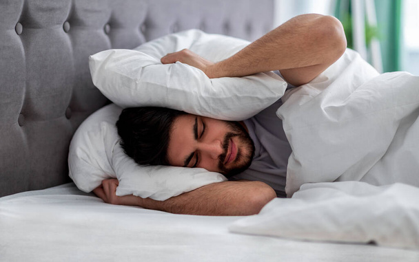 Rozrušený arabský muž si zacpává uši polštářem, skrývá se před příliš hlasitým zvukem nebo ranním hlukem, leží v posteli - Fotografie, Obrázek