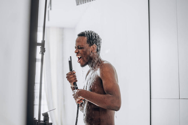 Szczęśliwy Afroamerykanin śpiewa pod prysznicem do wymyślonego mikrofonu pod strumieniem wody. - Zdjęcie, obraz