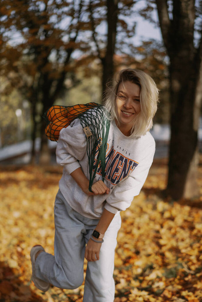 Young beautiful woman in a hat in an autumn park, a string bag with oranges, a woman throws up autumn leaves. Autumn mood, bright colors of nature. - Zdjęcie, obraz