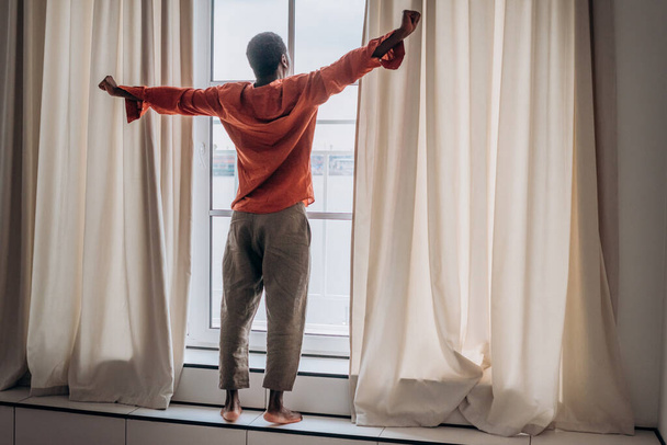 Az afrikai férfi nyújtózkodik, miután felébredt, az ablaknál állt és a távolba nézett. - Fotó, kép