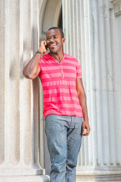 Kırmızı, pembe çizgili Henley V Neck tişörtü, gri pantolon, bilezik takan genç bir siyah adam bir sütunun yanında duruyor, gülümsüyor ve cep telefonuyla konuşuyor. - Fotoğraf, Görsel