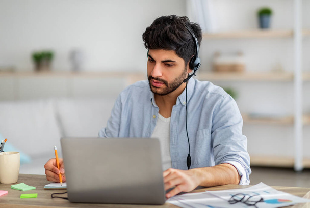 Hombre de negocios árabe feliz en los auriculares que tienen reunión de negocios en línea en el ordenador portátil, anotando información, sentado en la oficina en casa - Foto, imagen